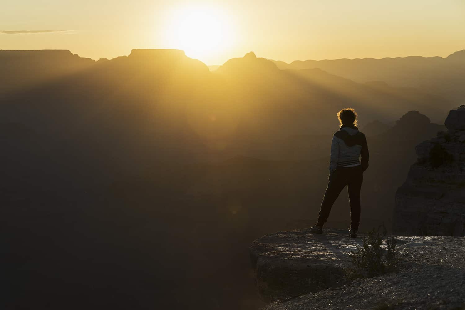A woman stands on a point overlooking a canyon at sunset.